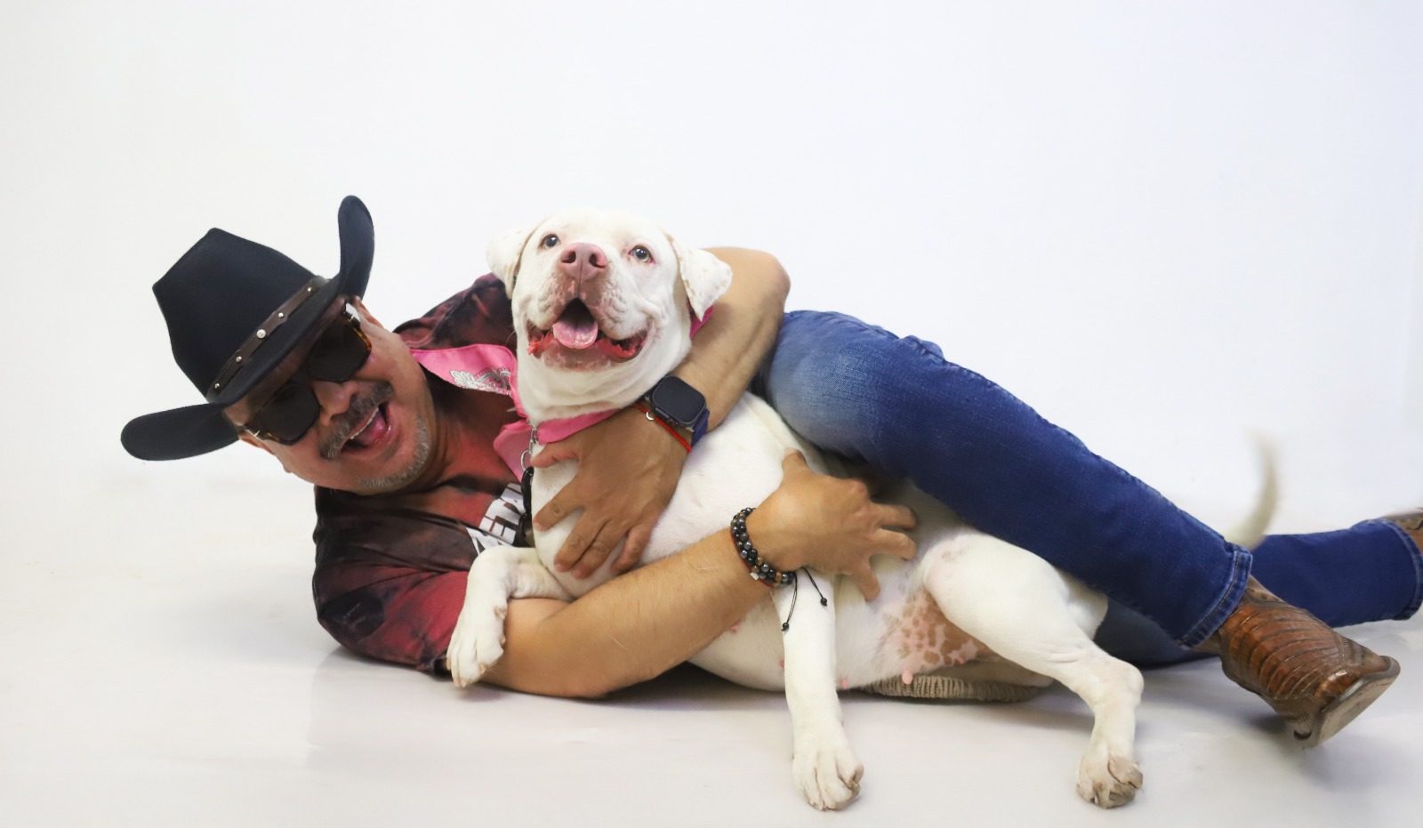 Hombre con sombrero y lentes abrazando a su perro blanco en el estudio.