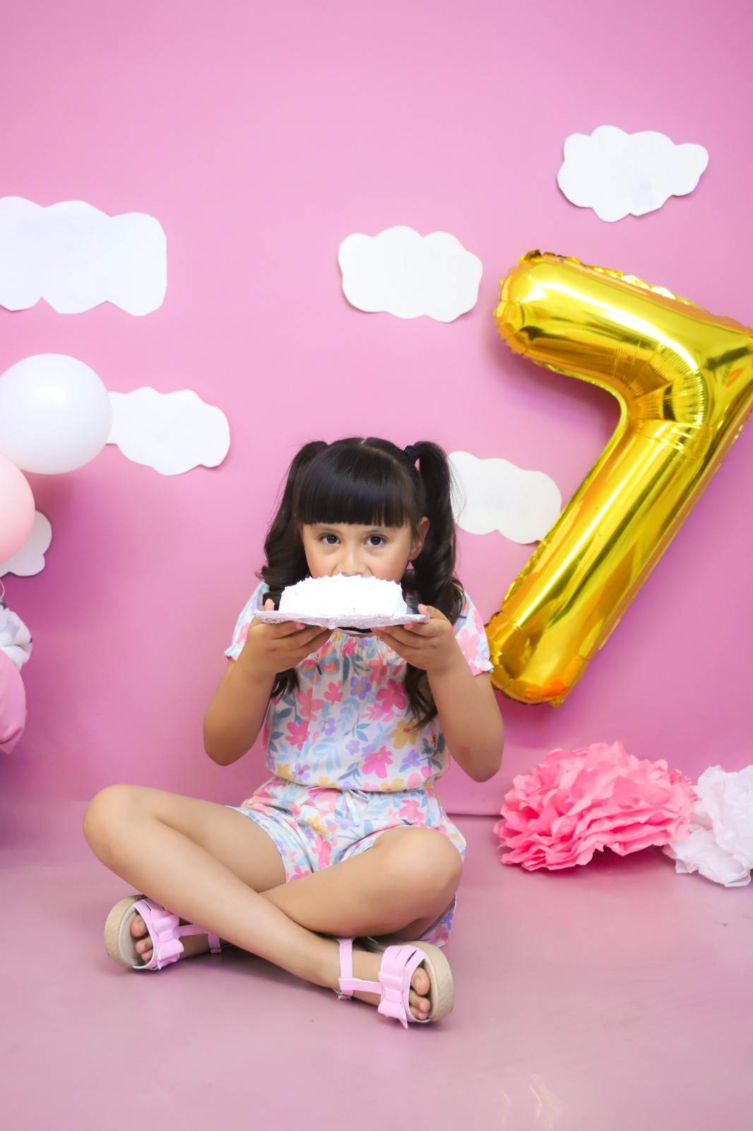 Niña comiendo pastel sentada frente a globos y decoración rosa.