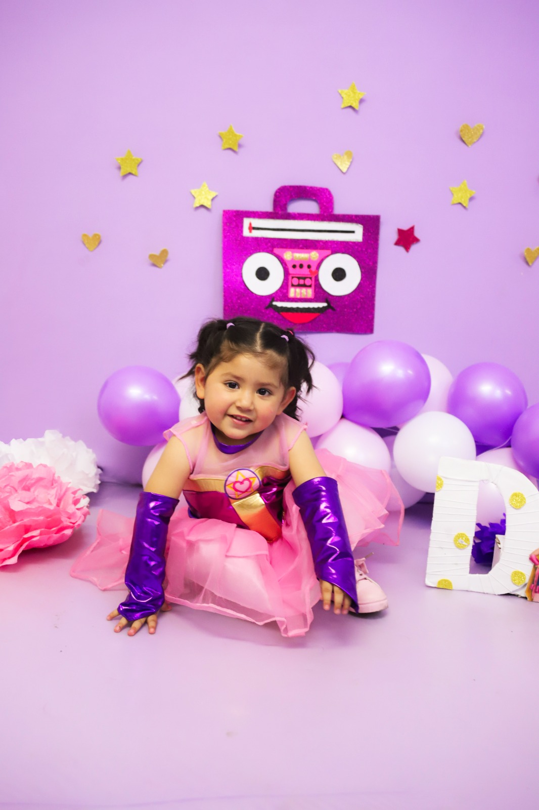 Niña con disfraz de princesa sentada entre globos morados.