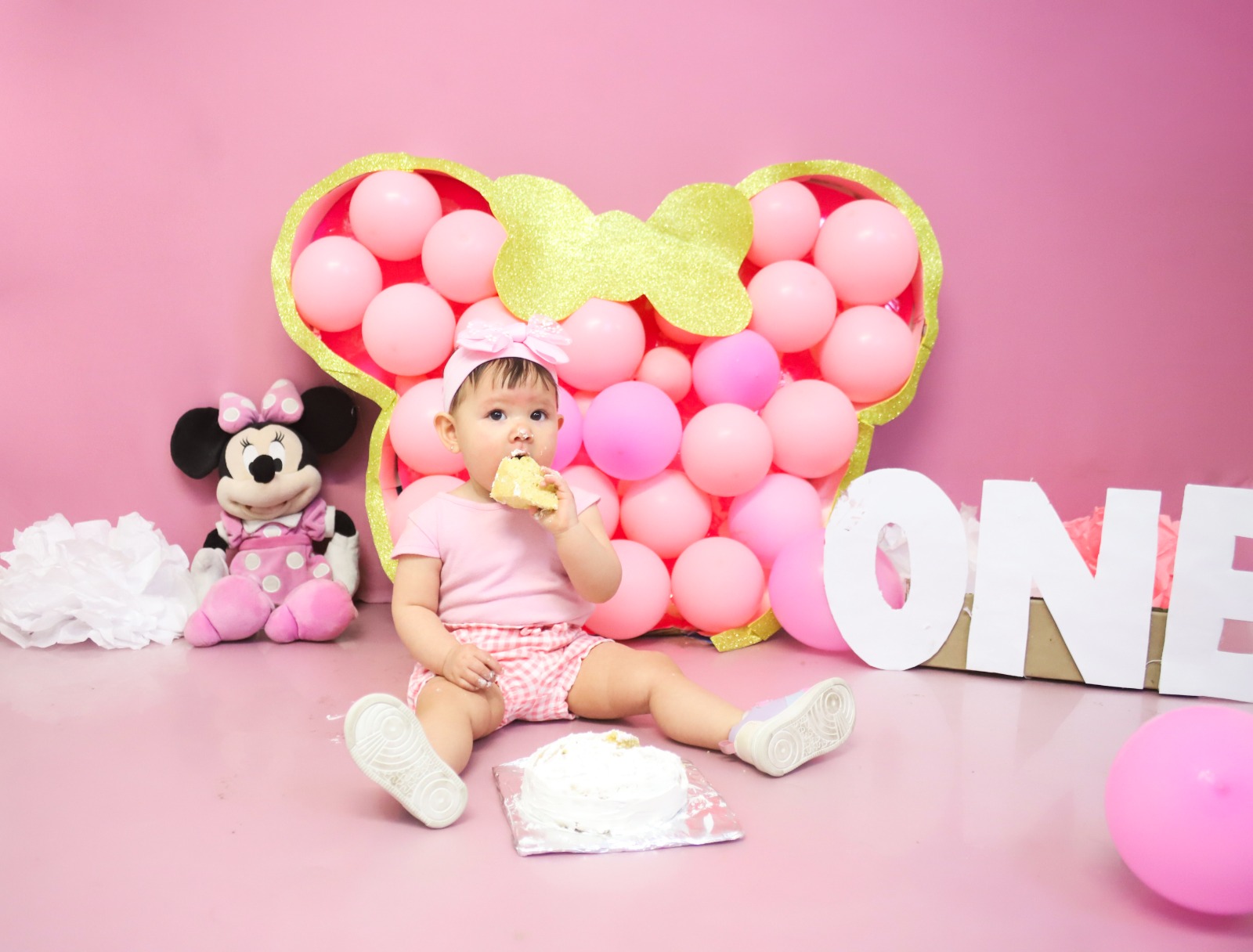 Bebé con diadema rosa comiendo un trozo de pastel frente a decoración de Minnie Mouse.