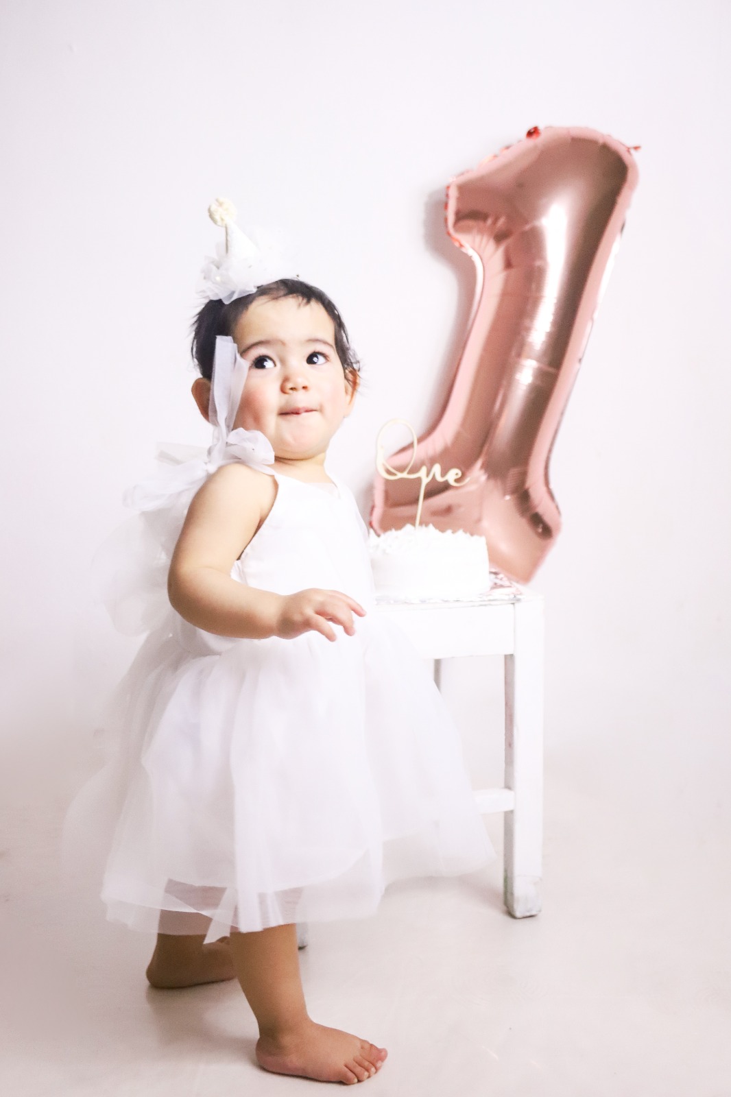 Bebé con vestido blanco junto al globo número uno y su pastel.